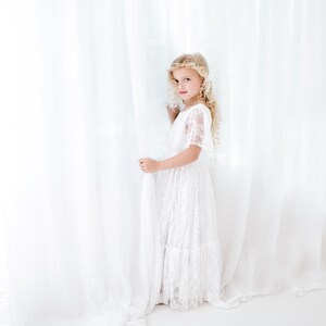 May include: A young girl wearing a white lace dress with a gold floral crown, standing in front of a white curtain.