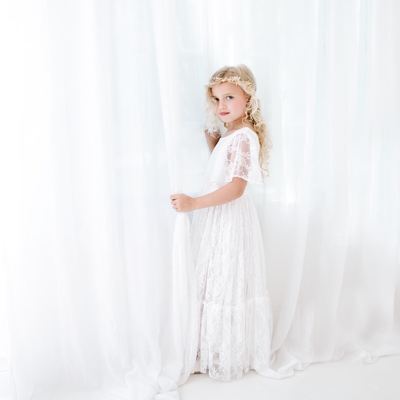 May include: A young girl wearing a white lace dress with a gold floral crown, standing in front of a white curtain.