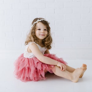 May include: A young girl wearing a white lace top and a pink tulle skirt sits on a white background. She is wearing a white flower crown and smiling at the camera.