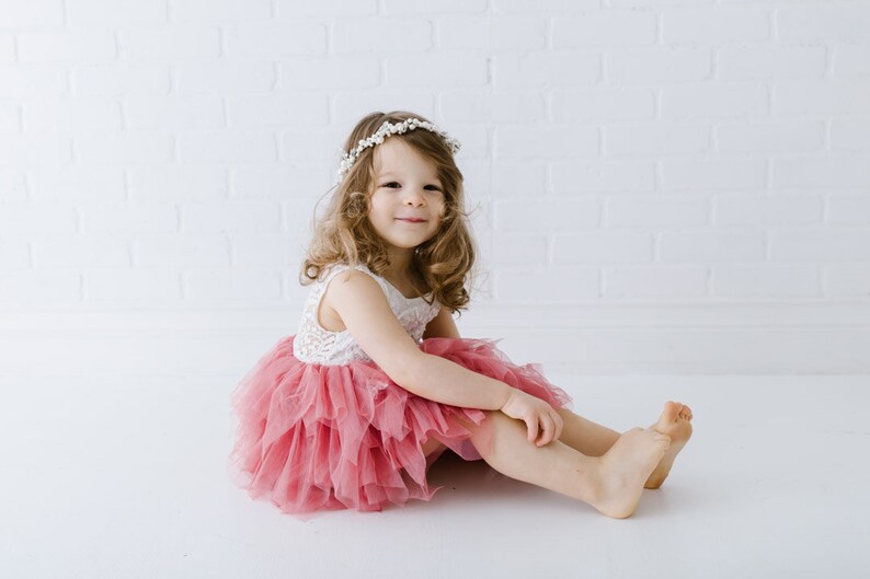 May include: A young girl wearing a white lace top and a pink tulle skirt sits on a white background. She is wearing a white flower crown and smiling at the camera.