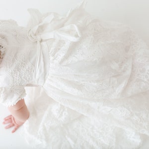 May include: A white lace dress with short sleeves and a tied ribbon bow at the waist. The garment features intricate floral lace patterns throughout the bodice and a full, flowing skirt. A matching white lace headband is worn on the head.