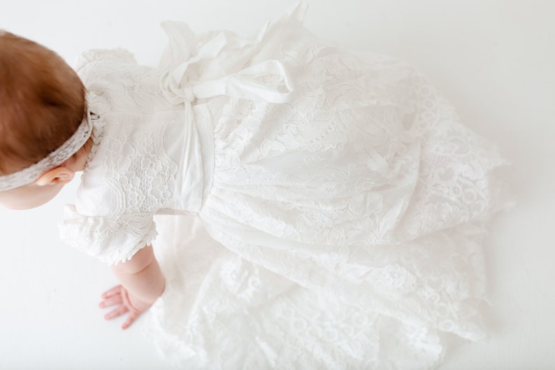 May include: A white lace dress with short sleeves and a tied ribbon bow at the waist. The garment features intricate floral lace patterns throughout the bodice and a full, flowing skirt. A matching white lace headband is worn on the head.