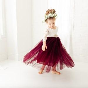 May include: A dress featuring a white lace bodice with three-quarter length sleeves and a full, floor-length skirt made of deep burgundy tulle. The waist is accented with a decorative, sparkling embellishment, and the skirt flares outward.