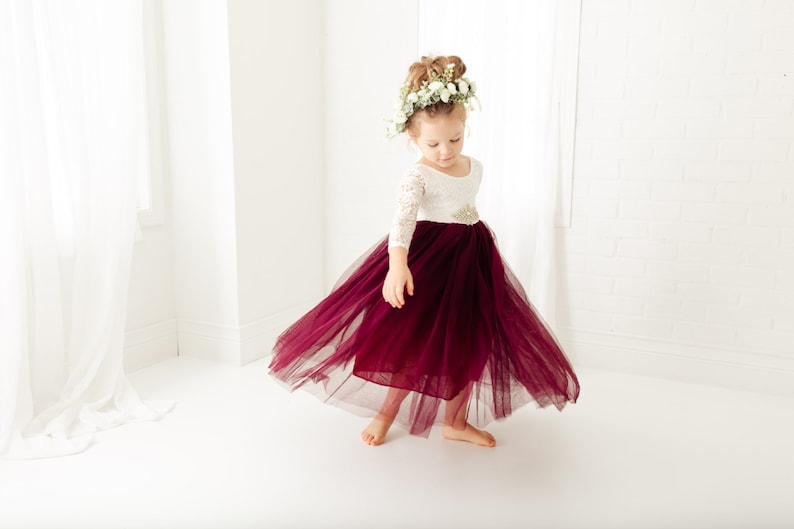 May include: A dress featuring a white lace bodice with three-quarter length sleeves and a full, floor-length skirt made of deep burgundy tulle. The waist is accented with a decorative, sparkling embellishment, and the skirt flares outward.
