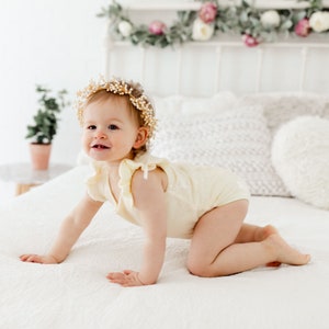 Puede incluir: Una ni&ntilde;a peque&ntilde;a con un body amarillo y una corona de flores est&aacute; gateando en una cama blanca.