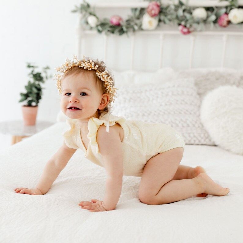 Puede incluir: Una ni&ntilde;a peque&ntilde;a con un body amarillo y una corona de flores est&aacute; gateando en una cama blanca.