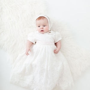 Puede incluir: Un beb&eacute; con un vestido de bautizo de encaje blanco y un gorro de crochet blanco. El beb&eacute; est&aacute; acostado sobre una superficie blanca y esponjosa.