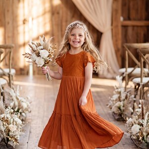 May include: A rust-colored dress with a lace bodice and short flutter sleeves, featuring a tiered, flowing skirt. The dress is paired with a floral headpiece and a bouquet of dried white flowers and grasses, complemented by light-colored ballet flats.
