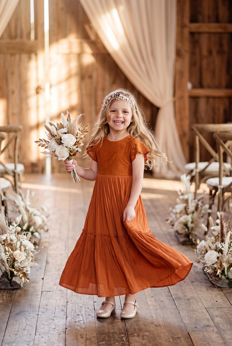 May include: A rust-colored dress with a lace bodice and short flutter sleeves, featuring a tiered, flowing skirt. The dress is paired with a floral headpiece and a bouquet of dried white flowers and grasses, complemented by light-colored ballet flats.