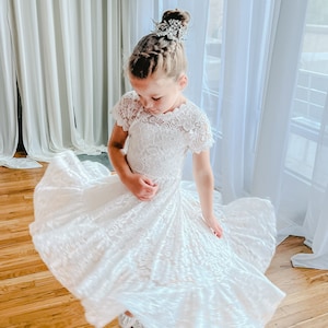 K&ouml;nnte beinhalten: Ein wei&szlig;es Spitzenkleid mit kurzen &Auml;rmeln und einem weiten, ausgestellten Rock, der eine Drehbewegung erzeugt. Das Kleid zeichnet sich durch ein zartes florales Spitzenmuster, einen gewellten Ausschnitt und einen ger&uuml;schten Saum aus. Ein silbernes Haaraccessoire wird getragen.