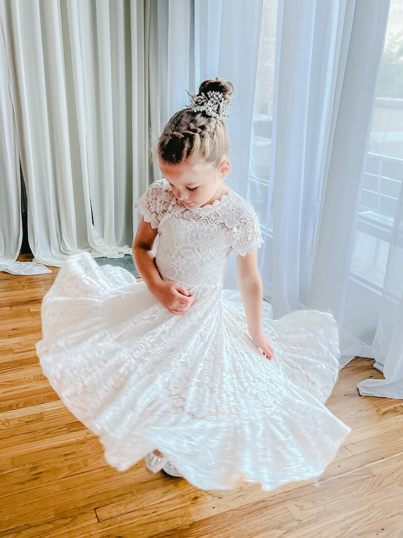 K&ouml;nnte beinhalten: Ein wei&szlig;es Spitzenkleid mit kurzen &Auml;rmeln und einem weiten, ausgestellten Rock, der eine Drehbewegung erzeugt. Das Kleid zeichnet sich durch ein zartes florales Spitzenmuster, einen gewellten Ausschnitt und einen ger&uuml;schten Saum aus. Ein silbernes Haaraccessoire wird getragen.