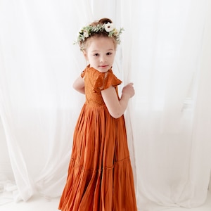 Op de afbeelding: Een jong meisje draagt een roestkleurige, gelaagde maxi-jurk met kantdetails en een bloemenkrans. De jurk heeft een aansluitend bovenlijf en een zwierige rok.