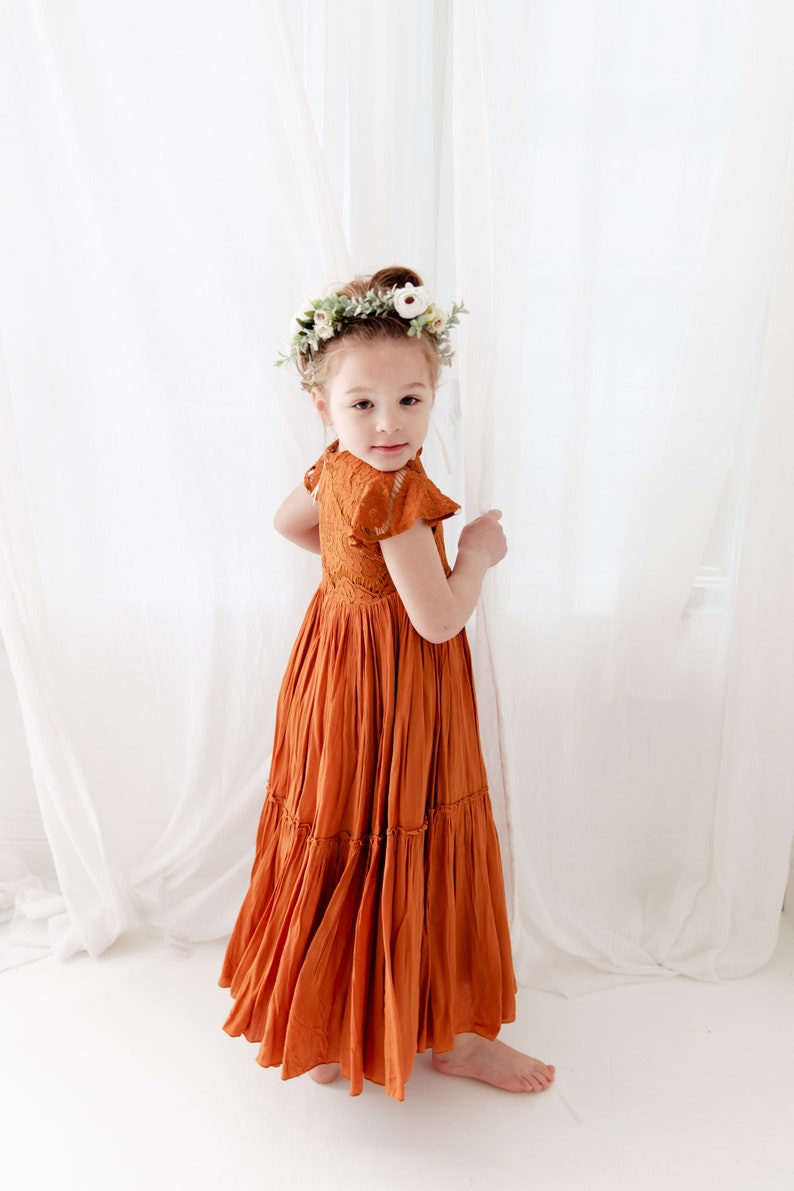 Op de afbeelding: Een jong meisje draagt een roestkleurige, gelaagde maxi-jurk met kantdetails en een bloemenkrans. De jurk heeft een aansluitend bovenlijf en een zwierige rok.