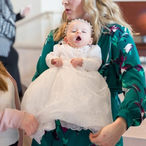 May include: A woman in a green floral dress holds a baby girl wearing a white lace christening gown. The baby is looking up and has a pearl headband on.