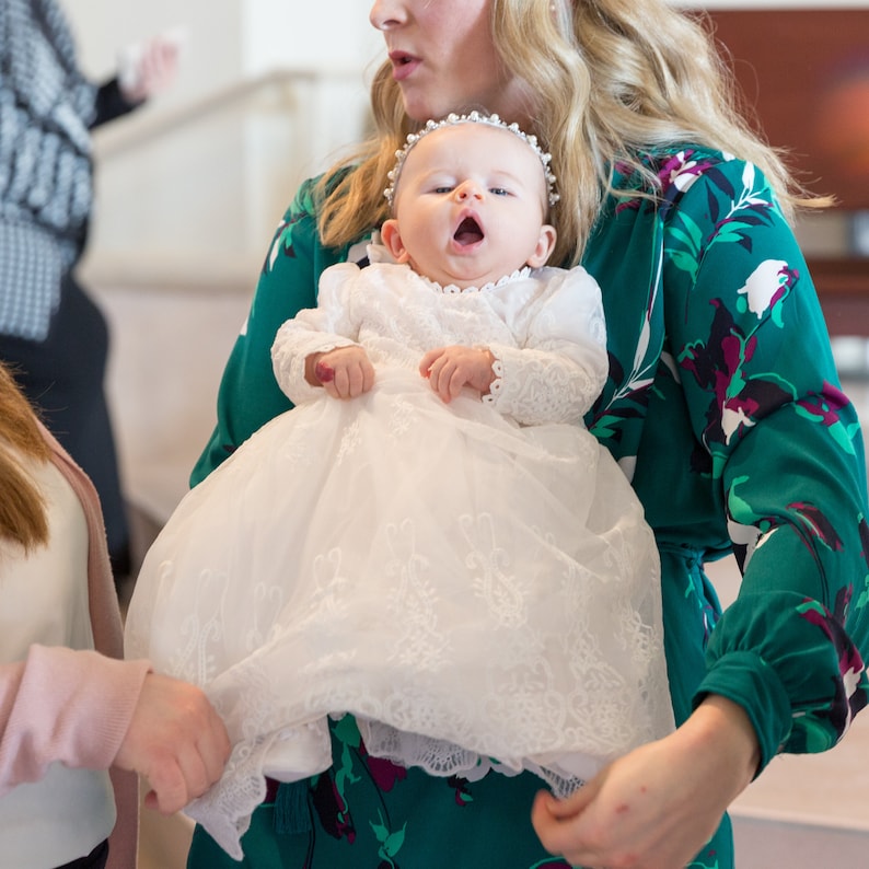 May include: A woman in a green floral dress holds a baby girl wearing a white lace christening gown. The baby is looking up and has a pearl headband on.