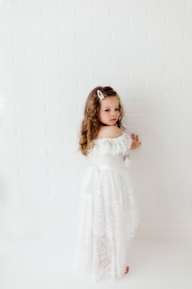 May include: A young girl wearing a white off-the-shoulder lace dress with a satin sash. She has curly brown hair and is wearing a pearl hair clip.