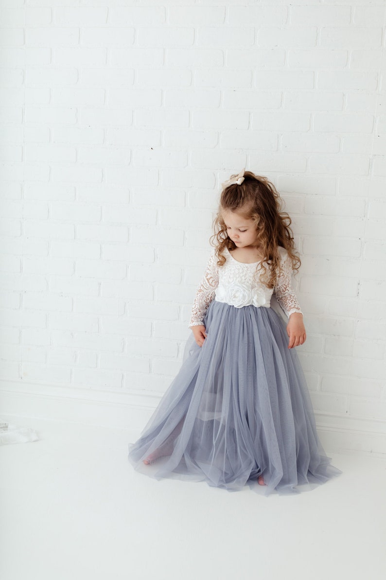 May include: A young girl wearing a white lace top with a tulle skirt in a light blue colour. The skirt is long and flowing, and the girl is standing in front of a white brick wall.