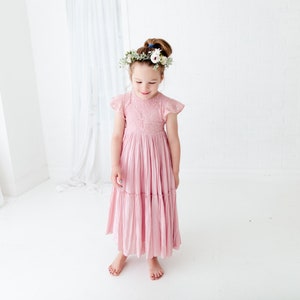 May include: A young girl wearing a pink, tiered, lace dress with a floral crown. The dress has short sleeves and a flowing skirt.
