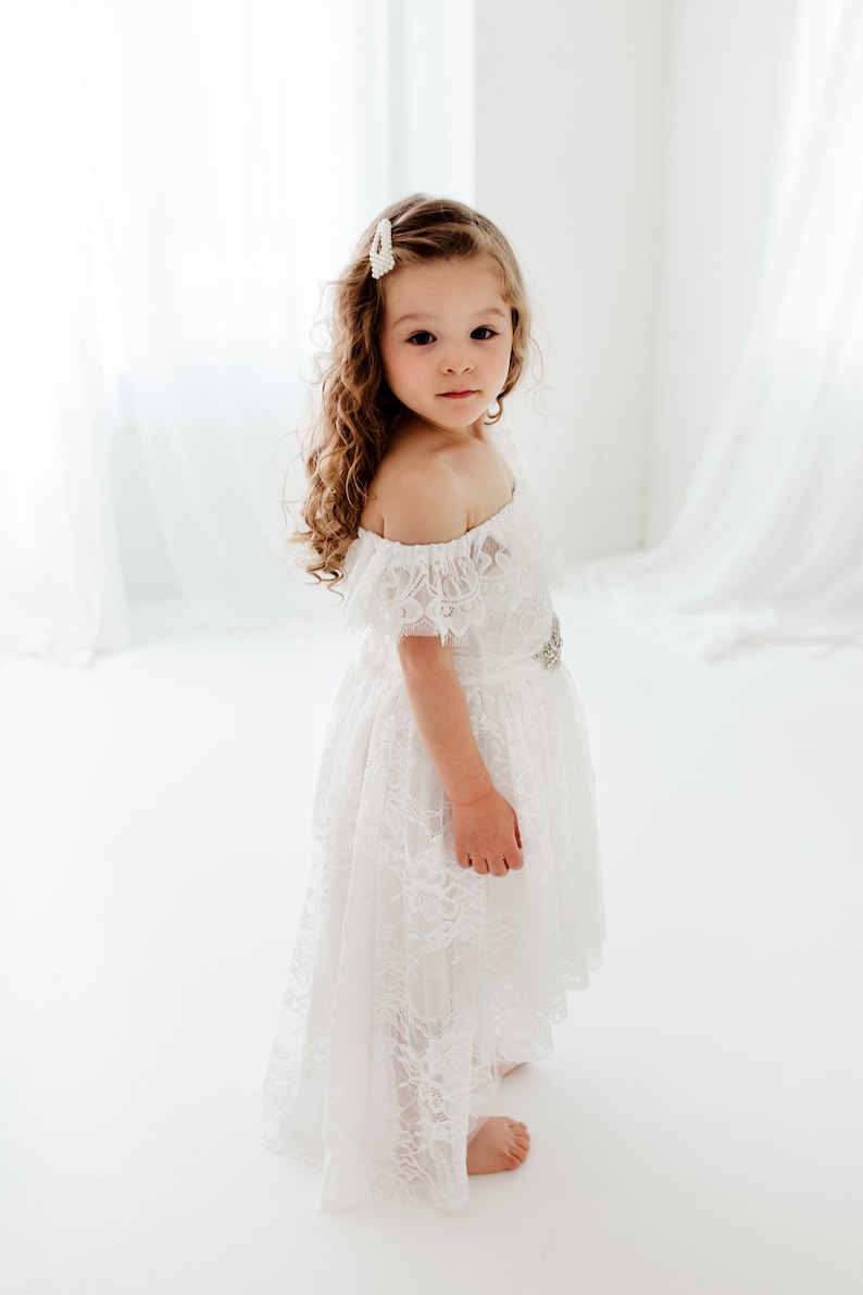 May include: A young girl wearing a white off-the-shoulder lace dress with a silver belt. She has long curly brown hair and is standing on a white background.