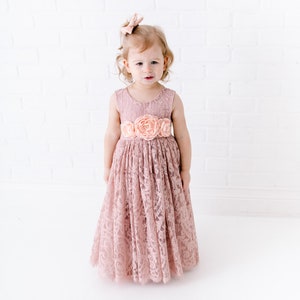 Pu&ograve; includere: Una bambina che indossa un abito di pizzo rosa polvere con una cintura di fiori abbinata. L'abito ha un corpetto senza maniche e una gonna ampia.