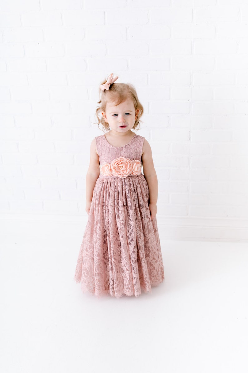 Pu&ograve; includere: Una bambina che indossa un abito di pizzo rosa polvere con una cintura di fiori abbinata. L'abito ha un corpetto senza maniche e una gonna ampia.