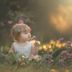 May include: A young child wearing a white dress with a pink tulle skirt sits in a field of purple flowers. The child is holding a single purple flower and looking off to the side.
