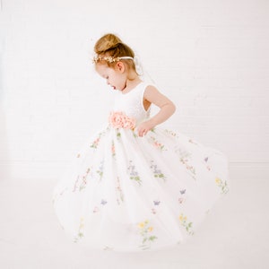 Puede incluir: Una ni&ntilde;a peque&ntilde;a con un vestido de tul blanco con estampado floral y una faja de flores de melocot&oacute;n. Lleva una diadema dorada con un adorno floral.