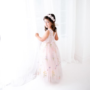 May include: A floor-length pale pink dress featuring a white lace bodice with a sleeveless design and a tulle skirt adorned with embroidered colourful wildflowers and small butterflies, paired with a matching floral headband with lace details.