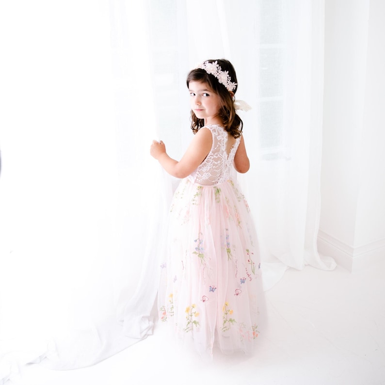 May include: A floor-length pale pink dress featuring a white lace bodice with a sleeveless design and a tulle skirt adorned with embroidered colourful wildflowers and small butterflies, paired with a matching floral headband with lace details.