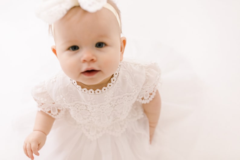 May include: A white dress featuring intricate lace detailing on the bodice, short puff sleeves, and a scalloped neckline. The garment includes a layered tulle skirt and is paired with a matching white floral headband accessory for a coordinated look.