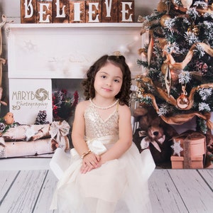 May include: A child wearing a sleeveless dress with a gold sequined bodice and a cream-colored tulle skirt, paired with a pearl necklace, a gold bow bracelet, and gold glittery ballet flats, sitting on a small white chair.