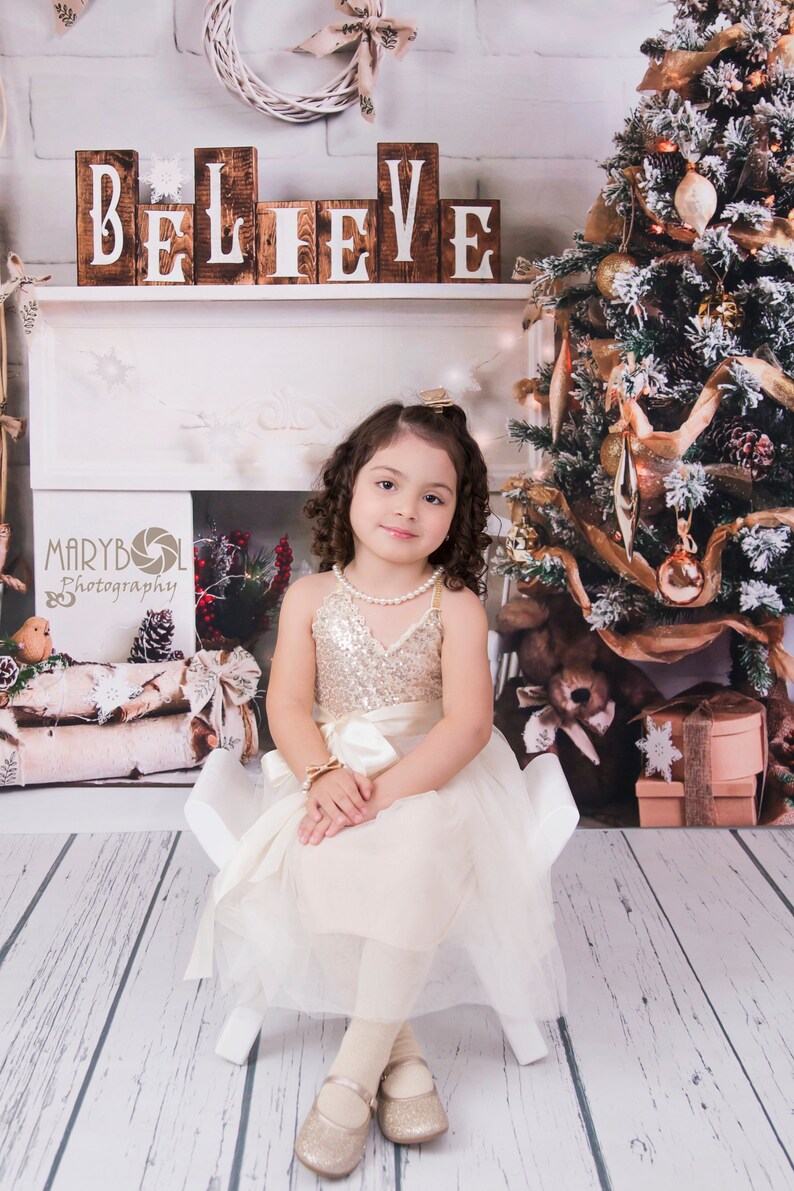 May include: A child wearing a sleeveless dress with a gold sequined bodice and a cream-colored tulle skirt, paired with a pearl necklace, a gold bow bracelet, and gold glittery ballet flats, sitting on a small white chair.