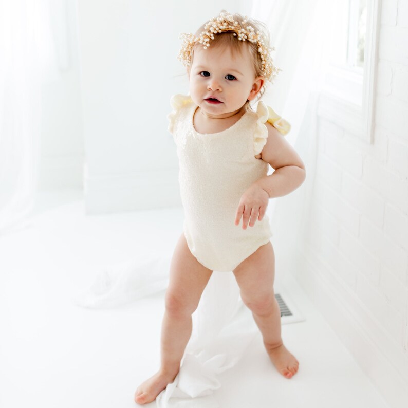 Puede incluir: Una ni&ntilde;a peque&ntilde;a con un body blanco con ribetes de volantes amarillos y una corona de flores. La beb&eacute; est&aacute; de pie sobre una superficie blanca y mirando a la c&aacute;mara.