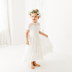 Op de afbeelding: Een witte kanten jurk met korte mouwen, een hoge halslijn en een rok tot op de grond. Het kledingstuk is voorzien van gedetailleerd bloemenkant over het lijfje en de rok. Een bloemenkrans met witte bloemen en groene bladeren wordt gedragen.