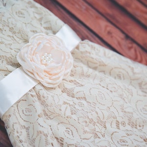 Peut inclure: Une robe en dentelle blanche avec un ruban en satin et une fleur en couleur p&ecirc;che.
