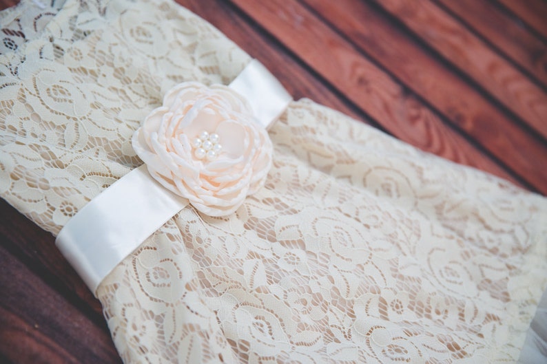 Peut inclure: Une robe en dentelle blanche avec un ruban en satin et une fleur en couleur p&ecirc;che.