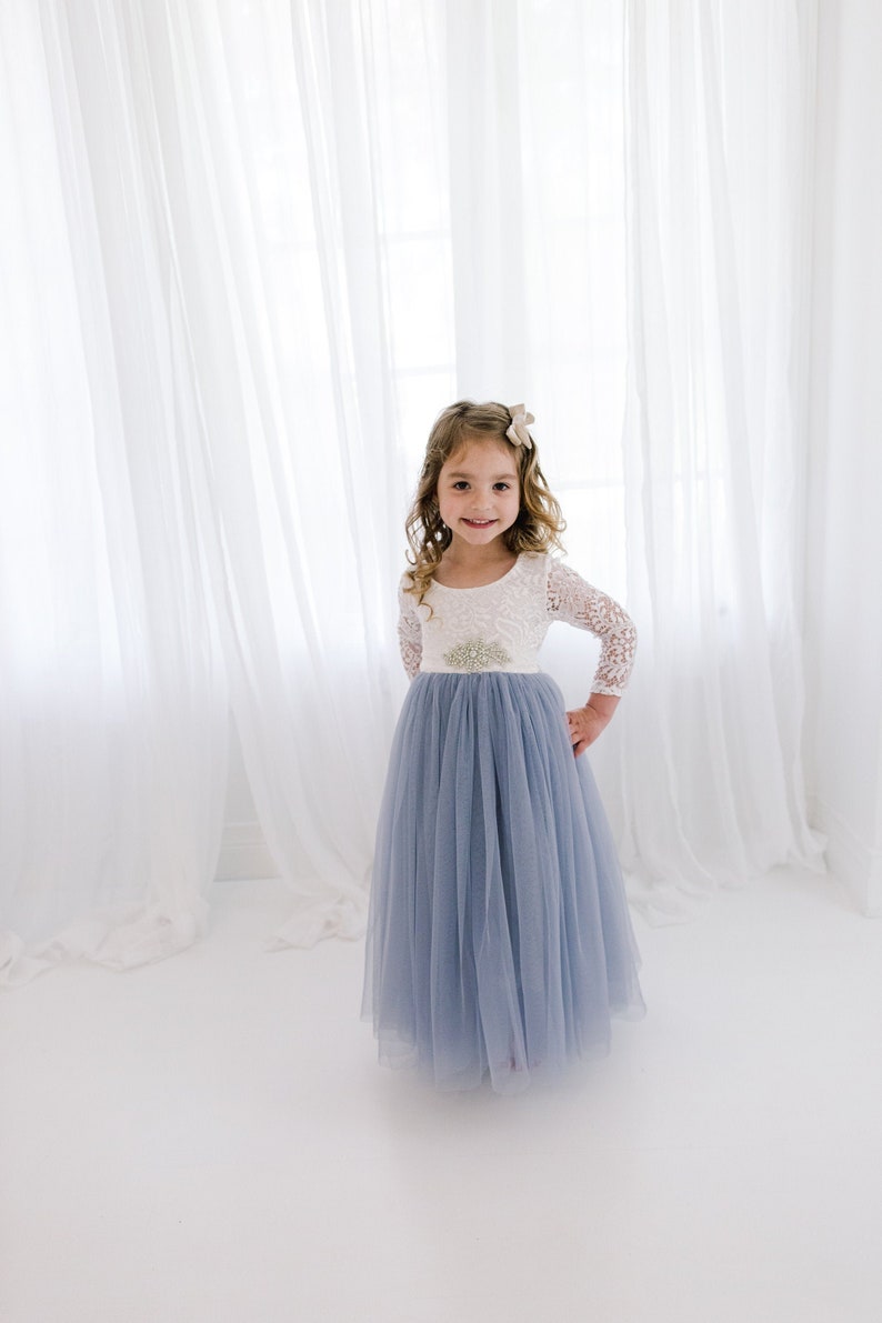 May include: A young girl wearing a light blue tulle dress with a white lace bodice and a silver belt. She is standing in front of a white curtain.