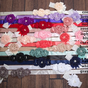 May include: A collection of 14 satin ribbon sashes with various colors and flower embellishments. The colors include cream, lavender, purple, red, rose, light pink, blush, coral, brown, mint, black, navy, white, and gray.