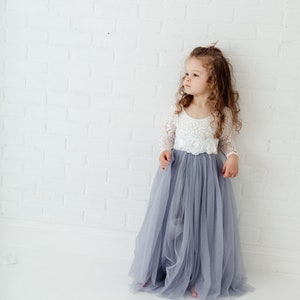 May include: A young girl wearing a white lace top with a long, flowing grey tulle skirt. She is standing in front of a white brick wall.