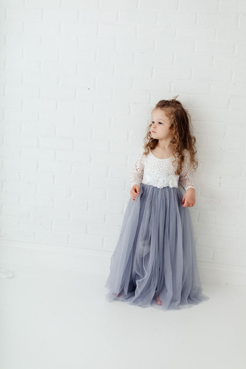 May include: A young girl wearing a white lace top with a long, flowing grey tulle skirt. She is standing in front of a white brick wall.