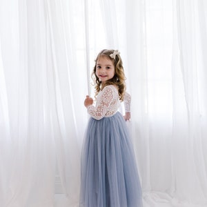 May include: A young girl in a white lace long-sleeved top and a light blue tulle skirt stands in front of a white curtain.