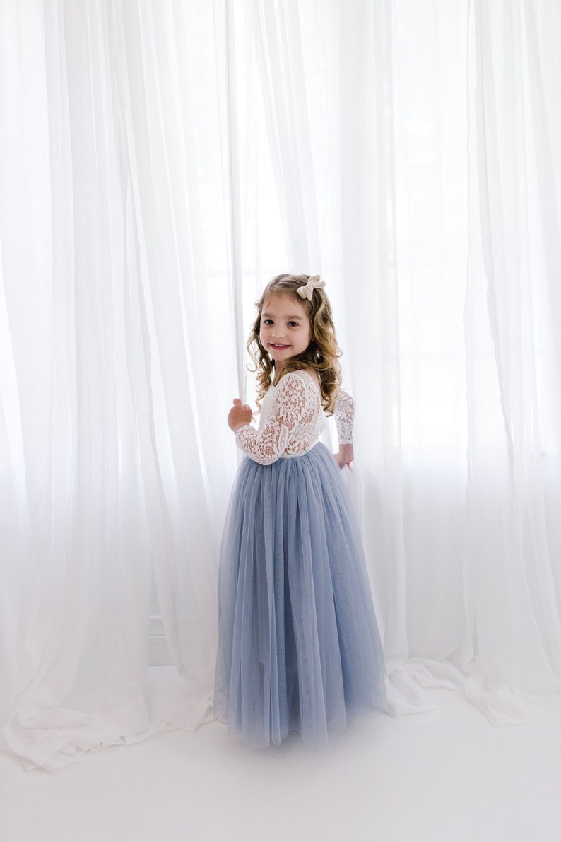 May include: A young girl in a white lace long-sleeved top and a light blue tulle skirt stands in front of a white curtain.