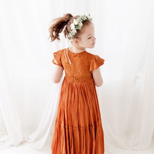 Op de afbeelding: Een jong meisje draagt een roestkleurige maxi-jurk met kantdetails en een witte bloemenkrans. De jurk heeft een gelaagde rok en korte mouwen.