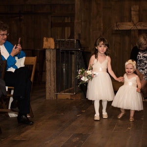 May include: Two young girls in white dresses walk down the aisle at a wedding ceremony. The girl in the front is holding a bouquet of flowers. The girl in the back is holding the hand of the girl in the front. The girls are walking towards a wooden cross.