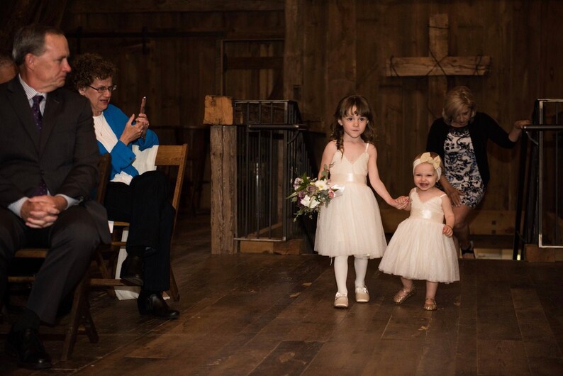 May include: Two young girls in white dresses walk down the aisle at a wedding ceremony. The girl in the front is holding a bouquet of flowers. The girl in the back is holding the hand of the girl in the front. The girls are walking towards a wooden cross.