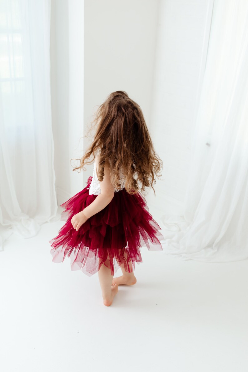 Puede incluir: Una ni&ntilde;a peque&ntilde;a con una parte superior de encaje blanco y una falda de tul burdeos en capas. La ni&ntilde;a se ve de espaldas, con cabello casta&ntilde;o largo y suelto. La falda tiene m&uacute;ltiples capas, creando una apariencia completa y esponjosa. El fondo es blanco brillante.