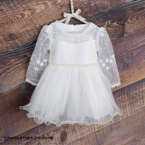 K&ouml;nnte beinhalten: Wei&szlig;es T&uuml;llkleid mit langen &Auml;rmeln, einer mit Perlen besetzten Taille und zarter Blumen Stickerei. Das Kleid h&auml;ngt an einem Holz Kleiderb&uuml;gel.