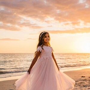 May include: A floor-length pale pink dress featuring a lace bodice with short sleeves and a full, layered tulle skirt. The garment is paired with a delicate floral headband, creating a cohesive look with a soft, flowing silhouette and intricate lace detailing.