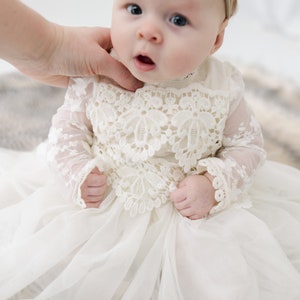 May include: An off-white infant dress featuring long sheer lace sleeves and a bodice with intricate floral lace applique. The skirt is made of layered, lightweight fabric that creates a full silhouette. The garment has a high neckline.