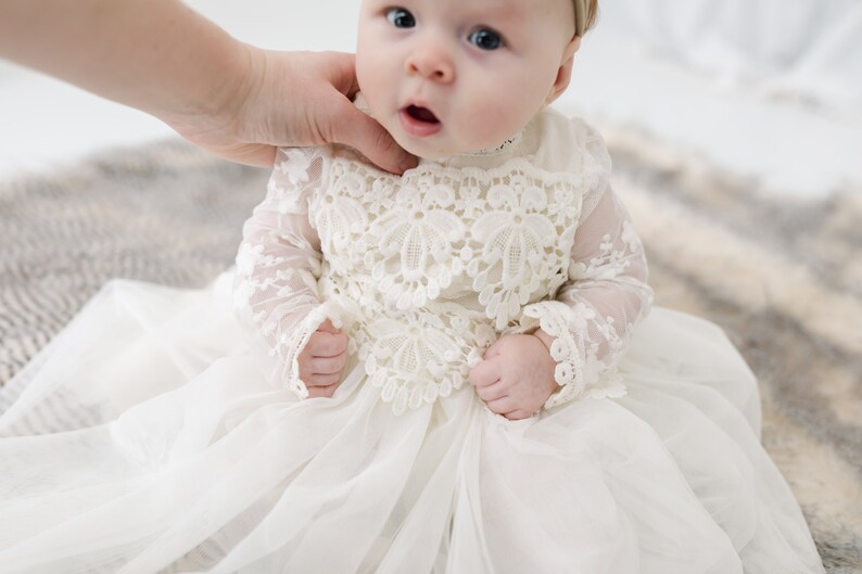 May include: An off-white infant dress featuring long sheer lace sleeves and a bodice with intricate floral lace applique. The skirt is made of layered, lightweight fabric that creates a full silhouette. The garment has a high neckline.